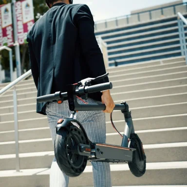 Teenage boy walking while holding a folded Hiboy S2 Pro electric scooter upright by its stem, with one hand gripping the handlebar.